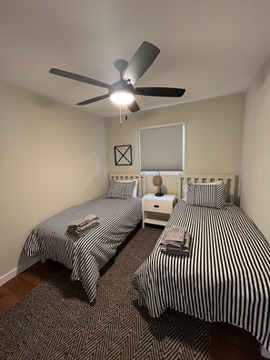 A bedroom with two single beds, a ceiling fan, a nightstand with a lamp, a window with blinds, and a wall-mounted picture. - River Perch on the Cacapon: private home on 27 acres and 1200 ft of riverfront (Yellow Spring)