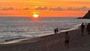 Escena de playa con el sol poniéndose sobre el océano, gente caminando por la orilla y un promontorio rocoso en la distancia.