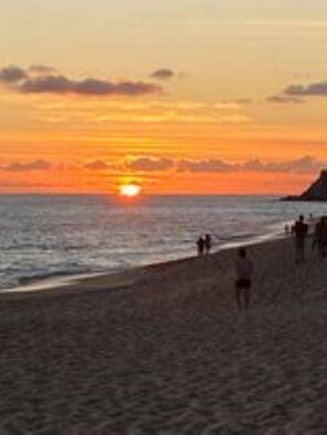 Une scène de plage au coucher du soleil sur l’océan, avec des personnes se promenant le long du rivage et un promontoire rocheux à l’horizon.