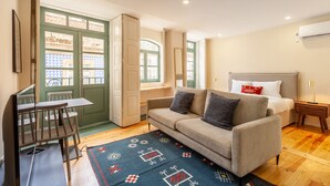 Une pièce confortable avec un canapé, une petite table et une chaise. On y trouve un lit avec des oreillers et une table de chevet. Une fenêtre offre une vue sur les bâtiments environnants.