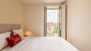 Une chambre d’hôtel avec un lit, un oreiller rouge, une table de chevet, une lampe et une vue sur les bâtiments à travers une porte ouverte.