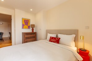 Un dormitorio con una cama grande, una mesita de noche con lámpara, una obra de arte en la pared y una puerta que da a otra habitación.