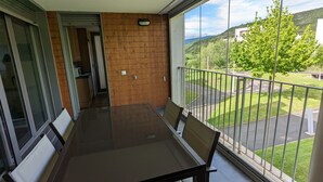Un balcon avec une porte-fenêtre donnant sur une cuisine, une table à manger avec des chaises, et une vue sur un paysage verdoyant avec des arbres et des bâtiments.