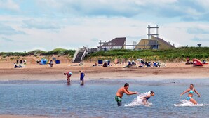 Une plage où des gens s’amusent dans l’eau, un escalier en bois menant à un bâtiment, ainsi que des chaises de plage et des parasols.