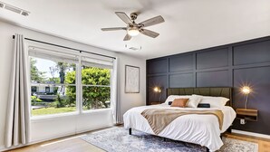 Ein Schlafzimmer mit großem Fenster, Deckenventilator, Bett mit weißer Bettwäsche und einer Wand in dunkler Farbe.