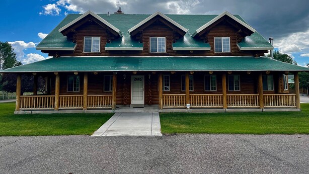 A two-story log cabin with a green roof, a porch with wooden railings, and a well-maintained lawn.