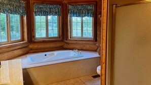 A log cabin bathroom with a large bathtub, wooden walls, and windows with floral curtains.