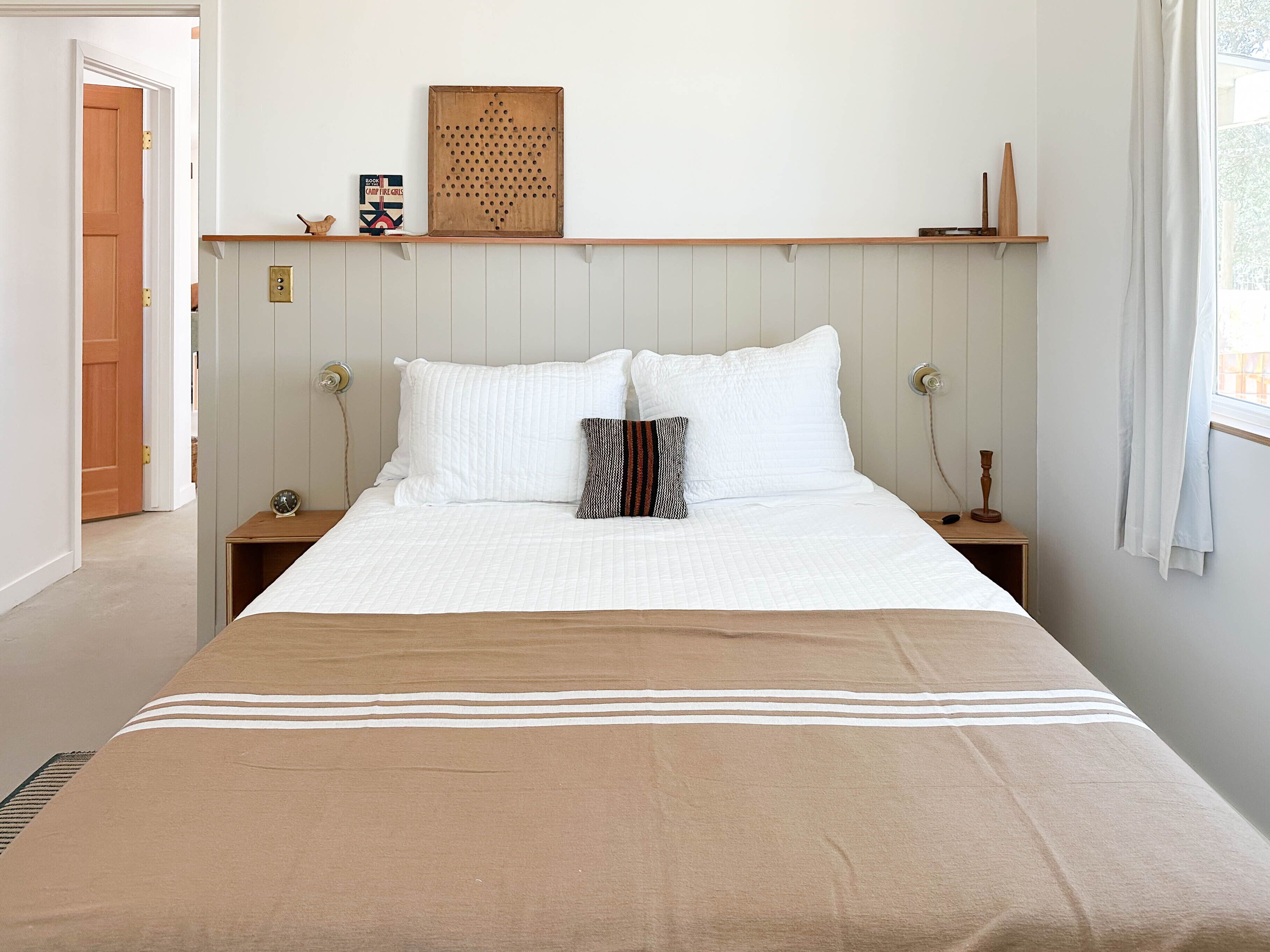 Una camera da letto con un letto, due comodini, una testiera in legno, una mensola a parete con oggetti decorativi e una finestra con tende.