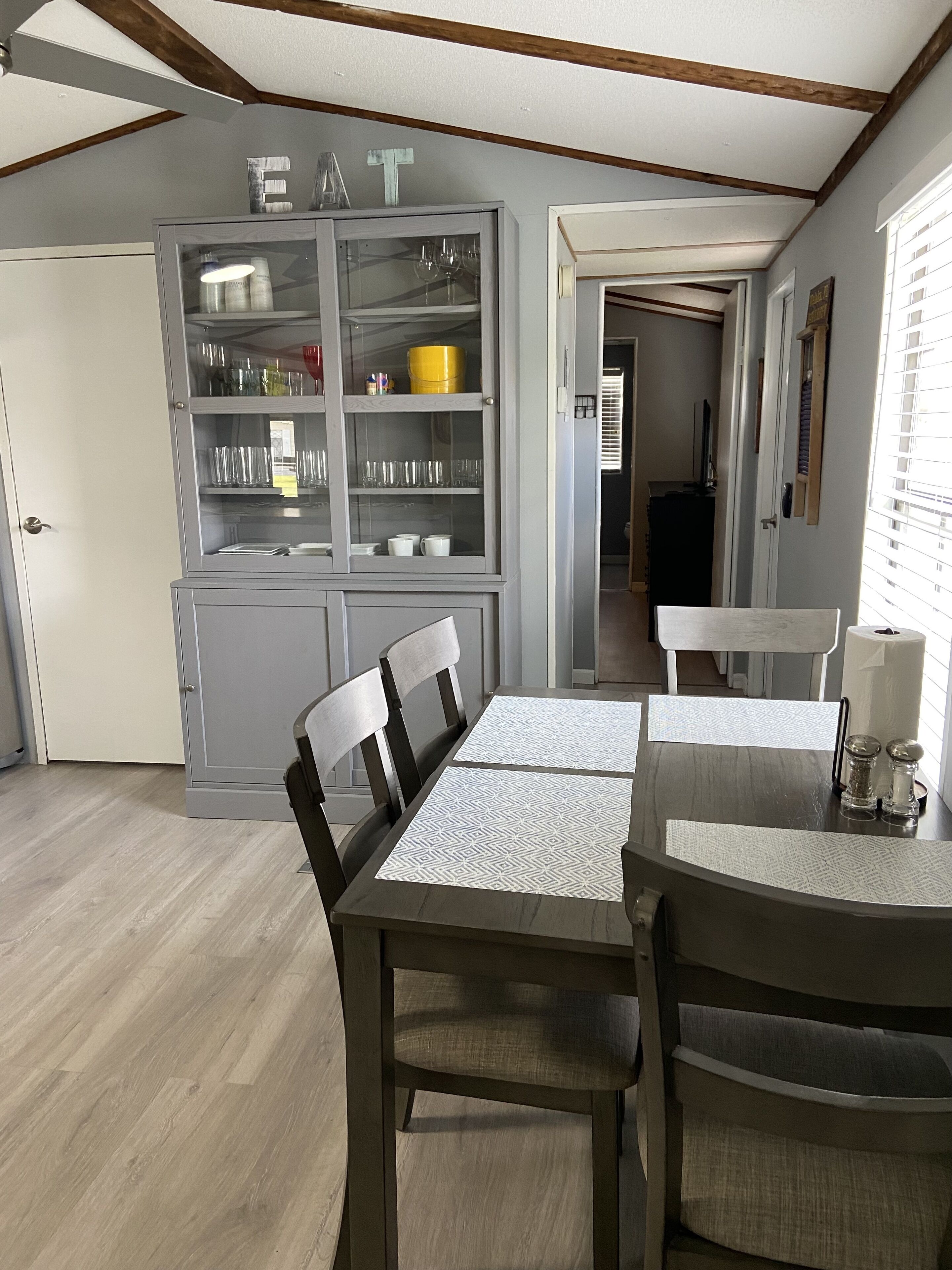 A dining area with a wooden table and chairs, a grey cabinet with glass doors containing kitchenware, and a view of the adjacent room through an open door.