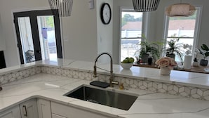 Une cuisine moderne avec un plan de travail en marbre blanc, un évier en acier inoxydable et des meubles blancs. On y trouve deux suspensions lumineuses, une horloge au mur et une fenêtre donnant sur l’extérieur.