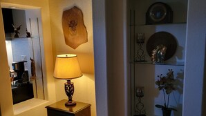 A room with a wooden floor, a lamp on a nightstand, a wall-mounted shelf with decorative items, and a mirror reflecting part of the room. - Saguaro National Park Westside retreat.   (Tucson)