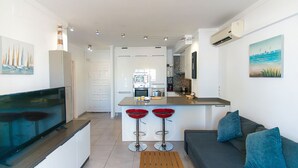 A modern kitchen with a central island, two red bar stools, a grey sofa with blue cushions, and a flat-screen TV on a white cabinet. - WELCS APARTAMENTO 059 EMP con vista al canal (Empuriabrava)