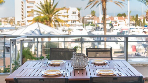 A dining table set for four with plates, cutlery, and a decorative vase, overlooking a marina with boats and palm trees. - WELCS APARTAMENTO 059 EMP con vista al canal (Empuriabrava)