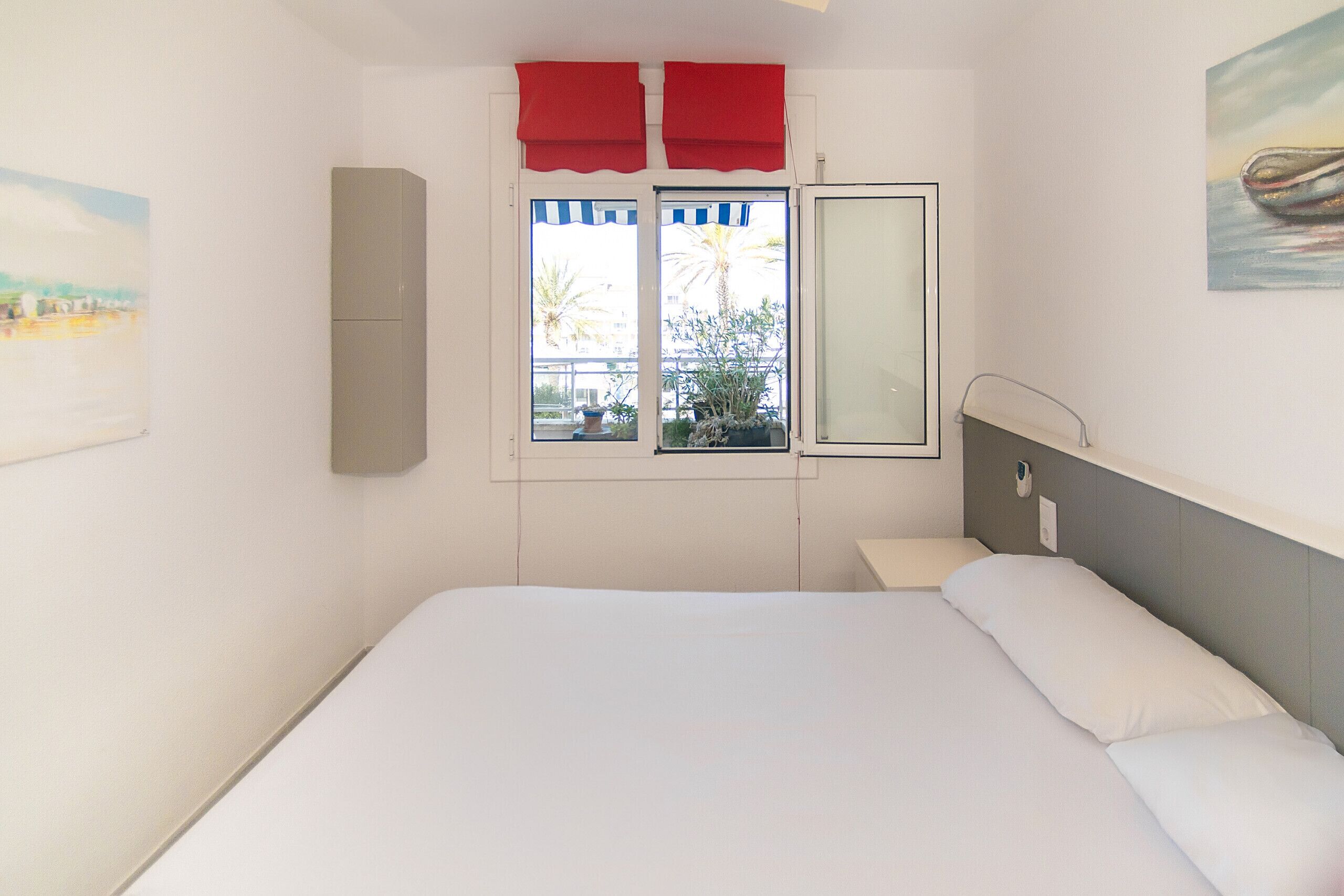 Un dormitorio con cama, ventana con cortinas rojas, mesita de noche y televisor instalado en la pared.
