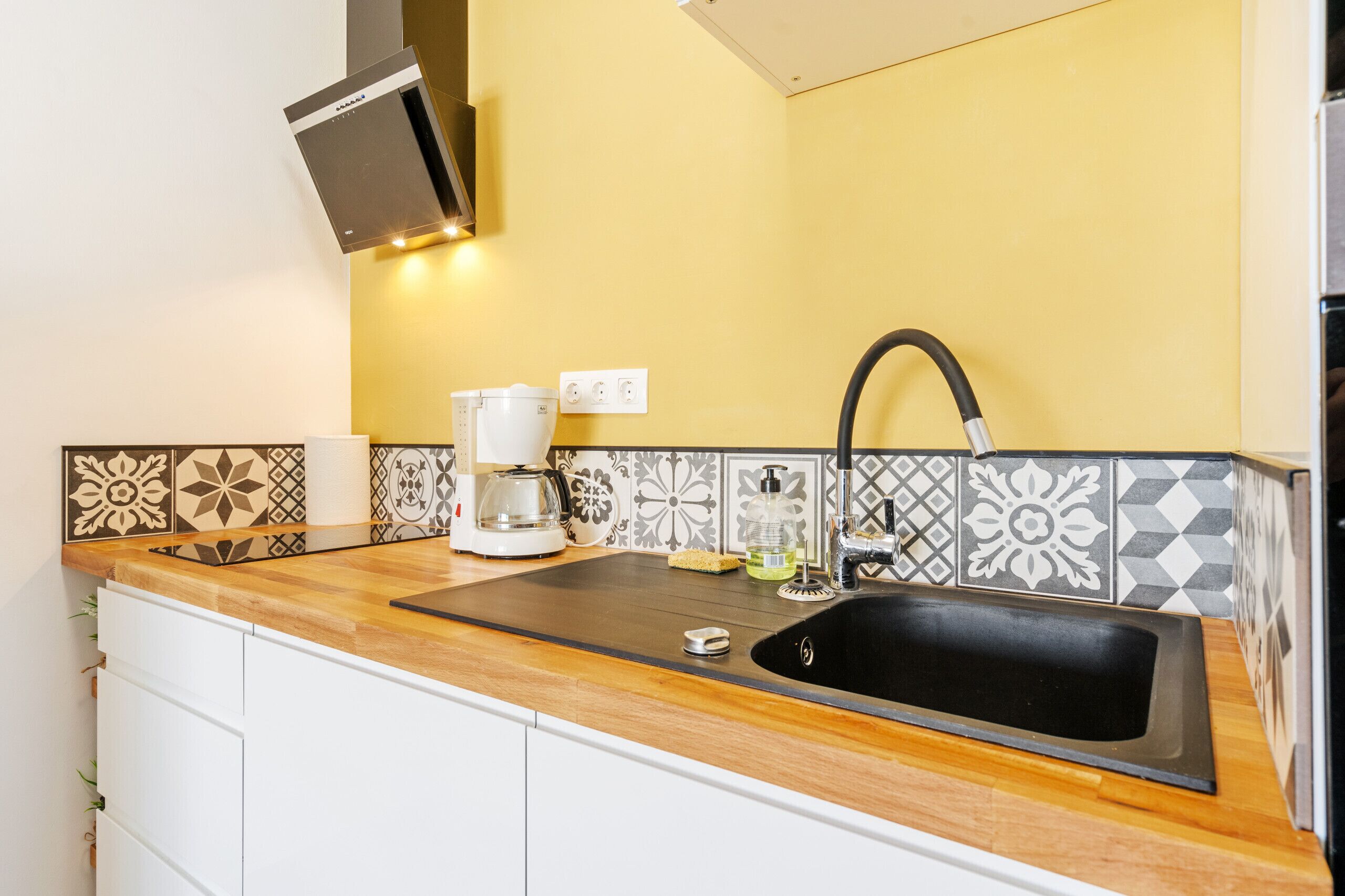A kitchen with a wooden countertop, a black sink, a coffee maker, and patterned backsplash tiles.
