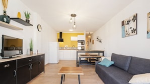A modern living room with a grey sofa, a wooden coffee table, and a TV on a black cabinet. There is a kitchen in the background with white cabinets and a yellow accent wall.