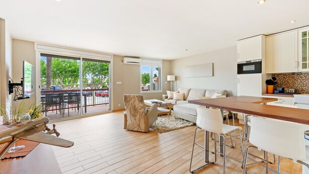 Uma sala de estar espaçosa com piso de madeira, uma grande porta de vidro deslizante que dá para um pátio externo, uma cozinha com armários brancos e um espaço bar com banquetas.