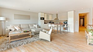 Uma sala de estar moderna com sofá, mesa de centro e um espaço de refeições com cadeiras, além de uma cozinha ao fundo.