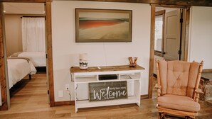 A room with a bed, a console table with a welcome sign, a chair, and a painting on the wall. - Old Chief Bunkhouse - ranch with views of Chief Mountain (Cardston)