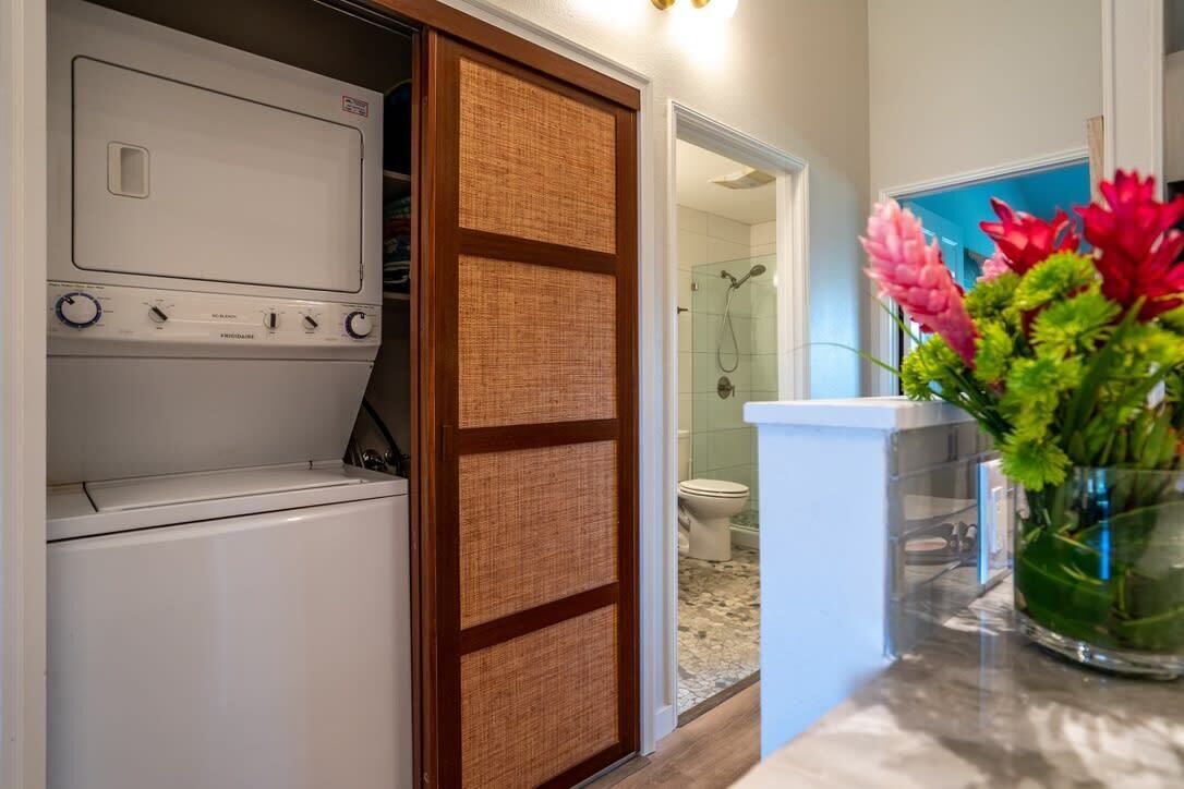 Une buanderie avec un lave-linge et un sèche-linge blancs, une porte coulissante en bois et une salle de bain visible à travers une porte ouverte.