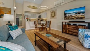 A modern living room with a sofa, coffee table, and TV. There is a kitchen in the background with a dining area. - Modern & Tropical Oceanside Vistas  (Kihei)