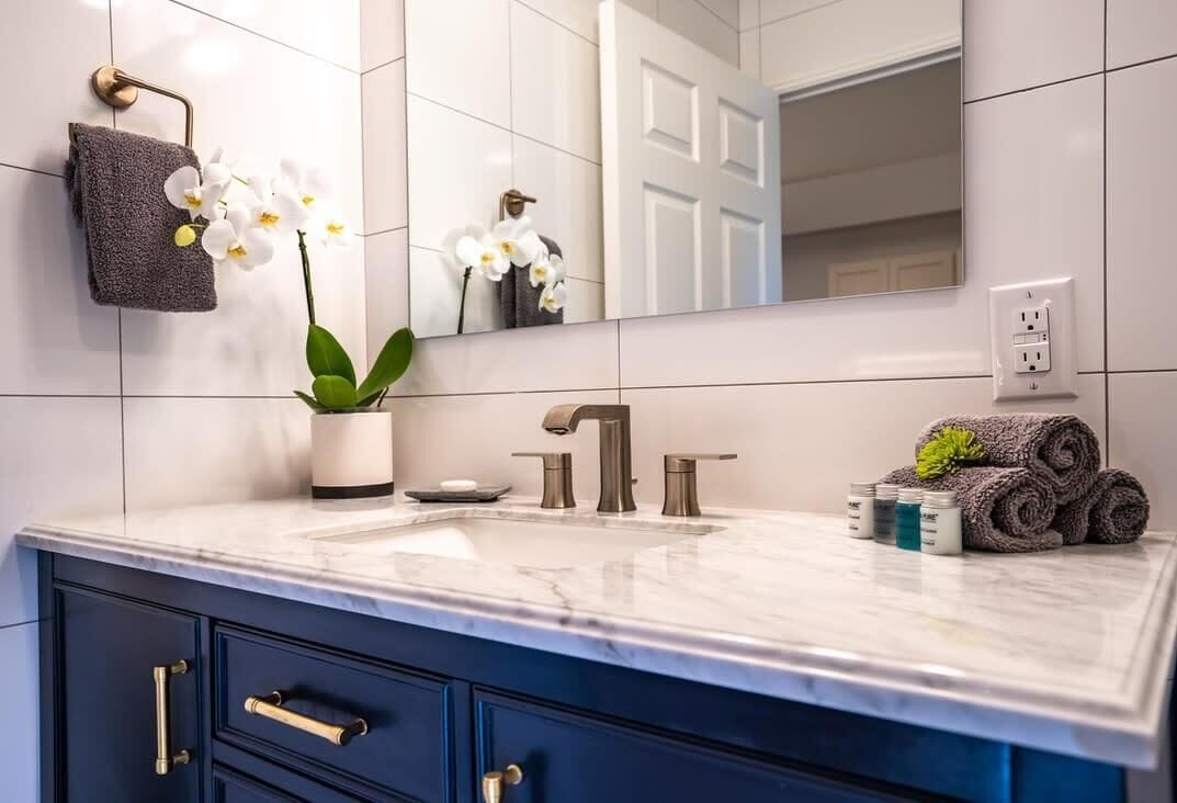 A bathroom with a marble sink, dark blue cabinets, a towel rack with a towel and orchid, and a mirror reflecting a door and wall.