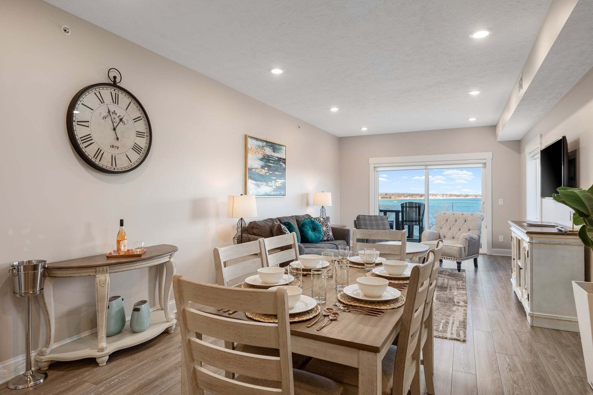A dining area with a round table set for four, a wall clock, and a view of the ocean through a large window.
