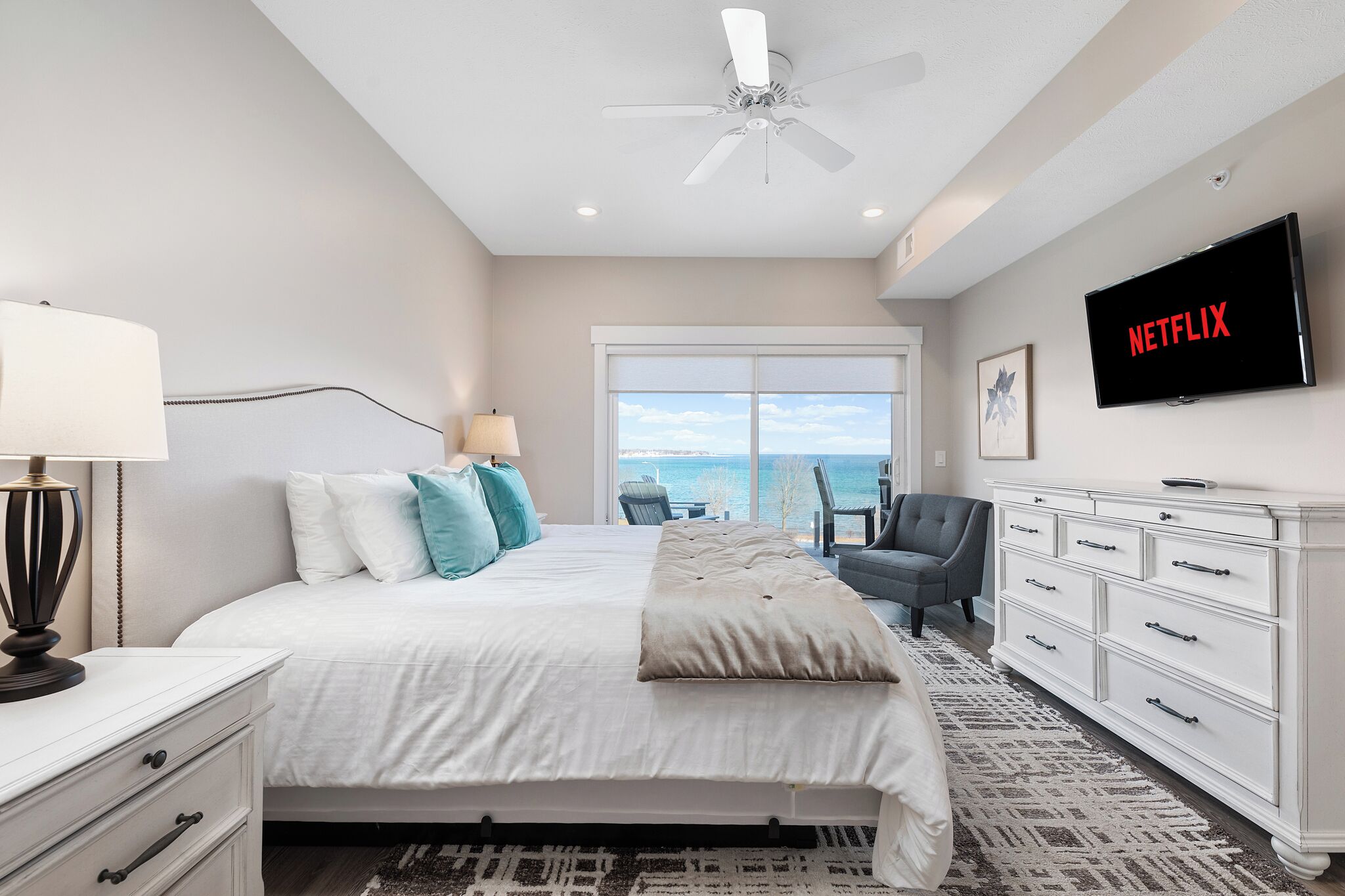 A bedroom with a bed, a TV, a ceiling fan, a dresser, and a view of the ocean.