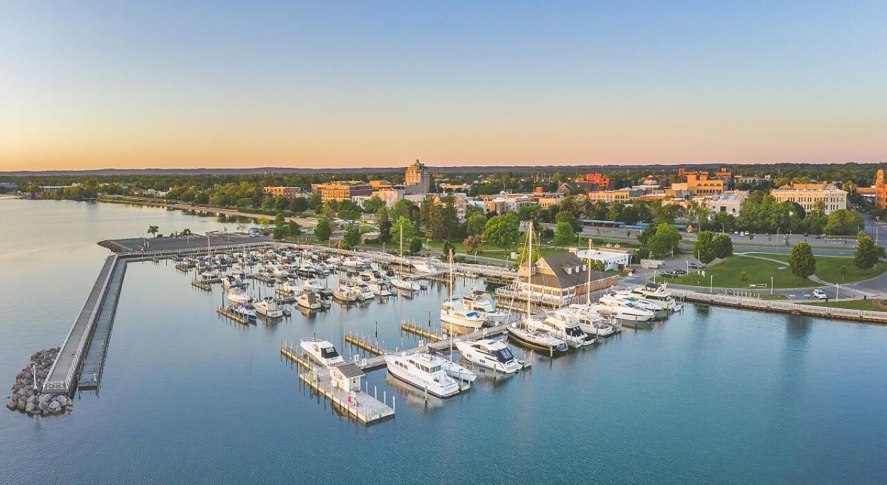 A marina with boats docked, a pier extending into the water, and buildings in the background.