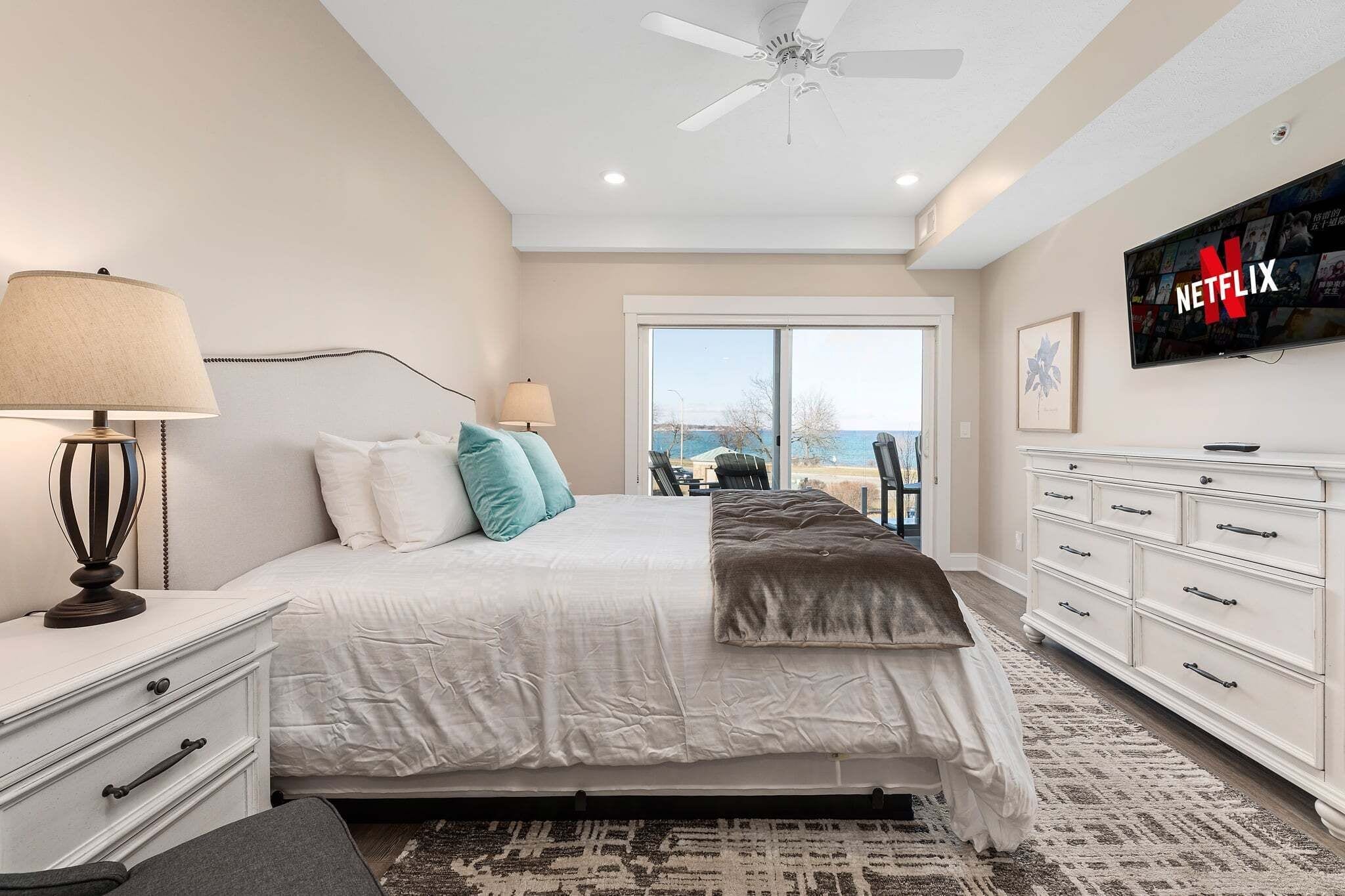 A bedroom with a bed, bedside tables, a TV, a ceiling fan, and a view of the outdoors.