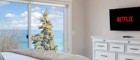 A bedroom with a bed, a dresser, a TV, and a view of the sea and trees.