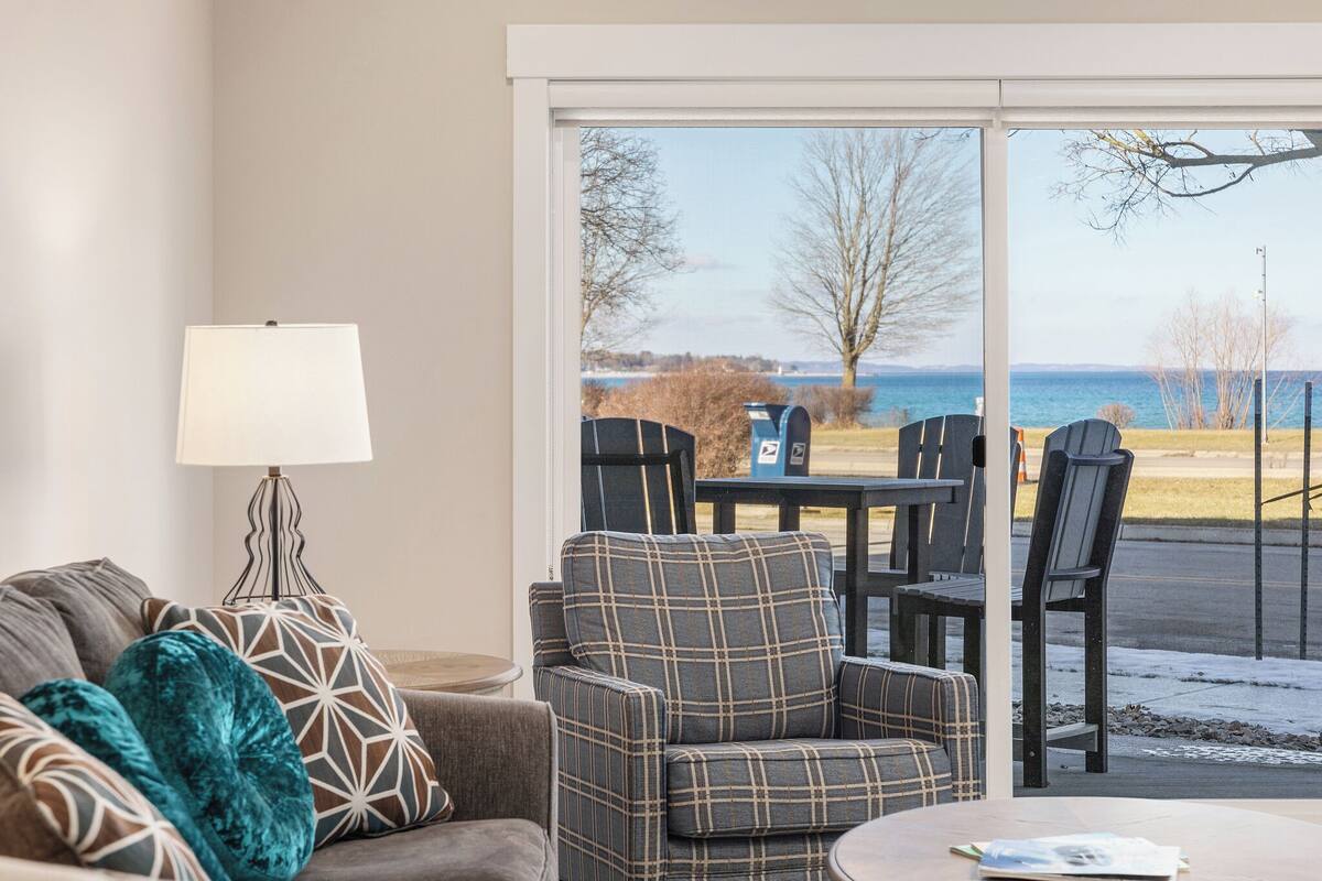 A living room with a sofa, armchair, and a table with chairs, a lamp, and a view of the outdoors.
