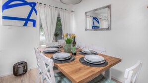 Uma área de refeições com uma mesa de madeira posta para quatro pessoas, cadeiras brancas, um vaso com flores e uma garrafa de vinho.
