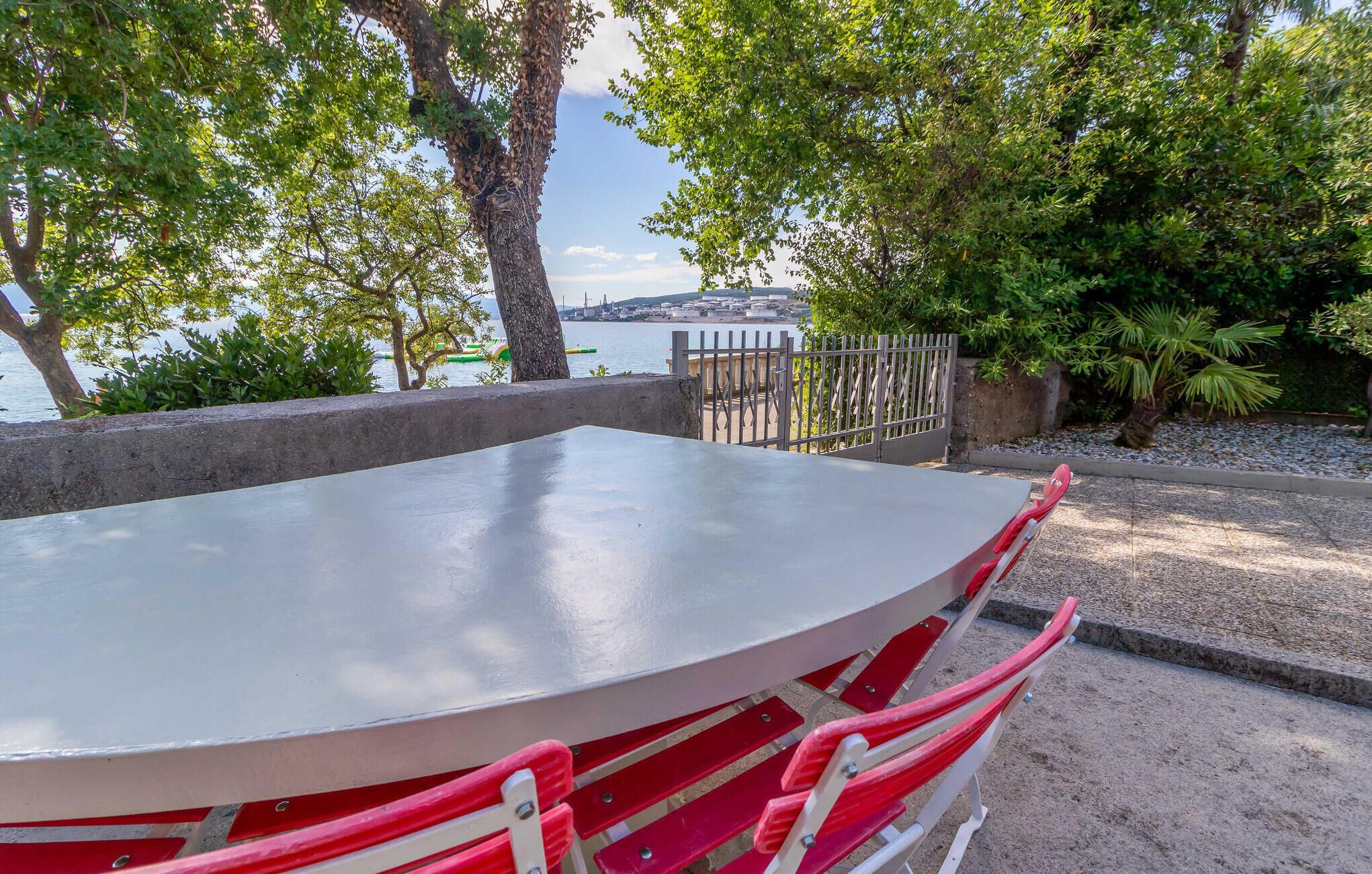 A table with red chairs, a concrete barrier, and a view of a body of water with buildings in the background.