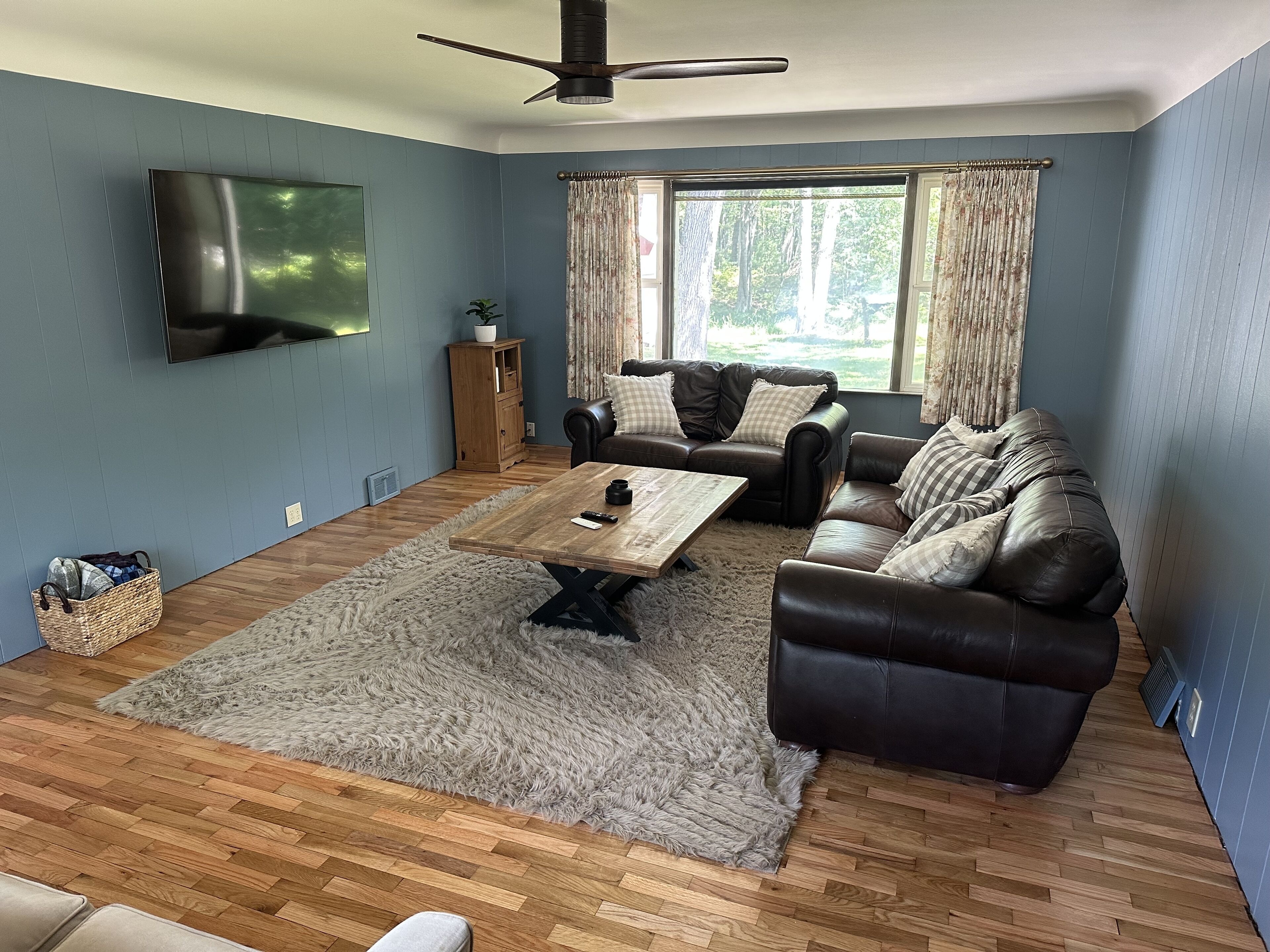 Una sala de estar con una mesa de centro de madera, dos sofás de cuero, un televisor montado en la pared y una ventana con cortinas.