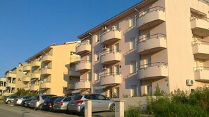 Une rangée d’immeubles à appartements de plusieurs étages, dotés de balcons, avec des voitures garées et un ciel dégagé.
