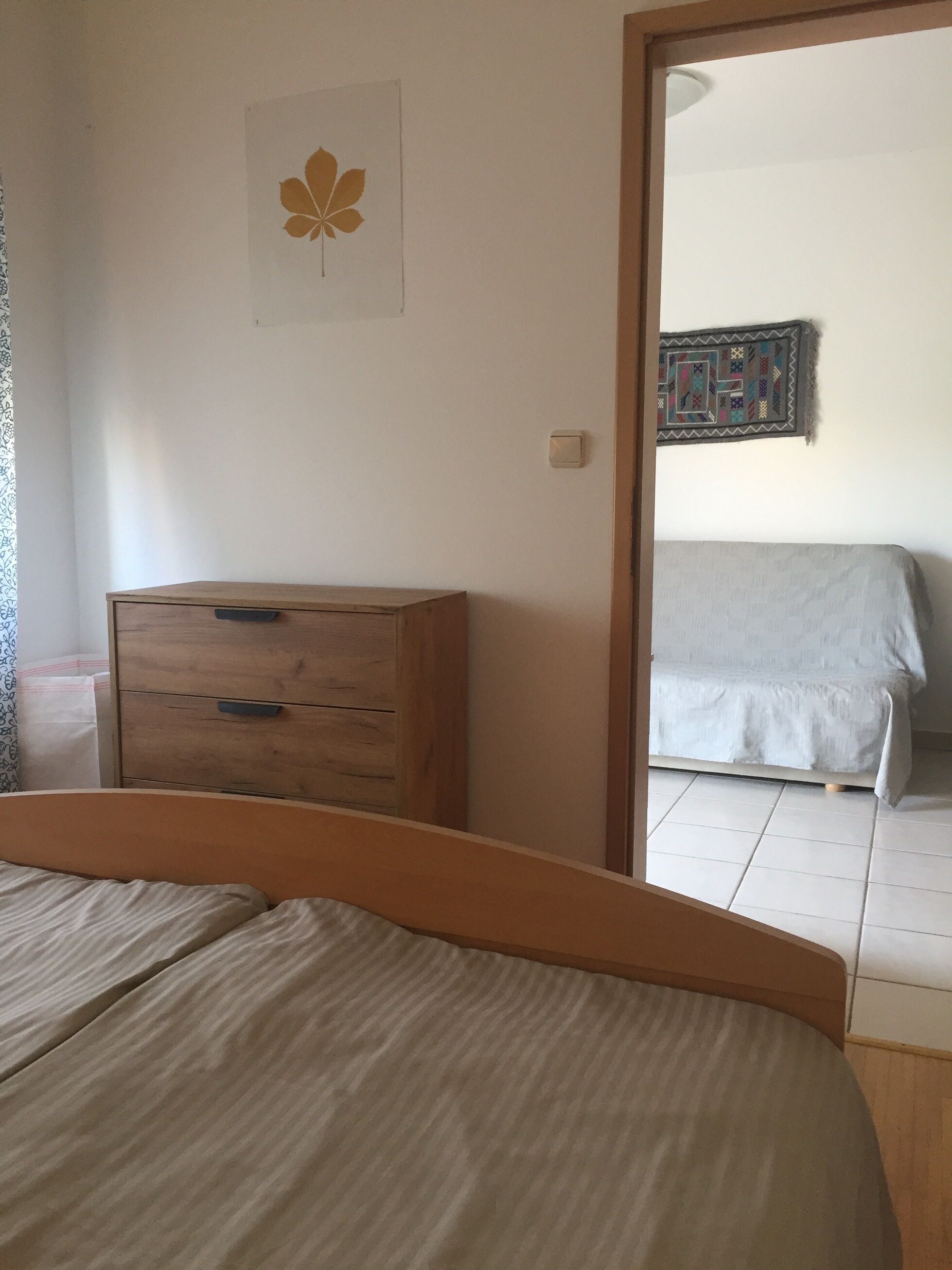 A bedroom with a wooden dresser, a bed with a white bedspread, a wall-mounted quilt, and a door leading to another room.