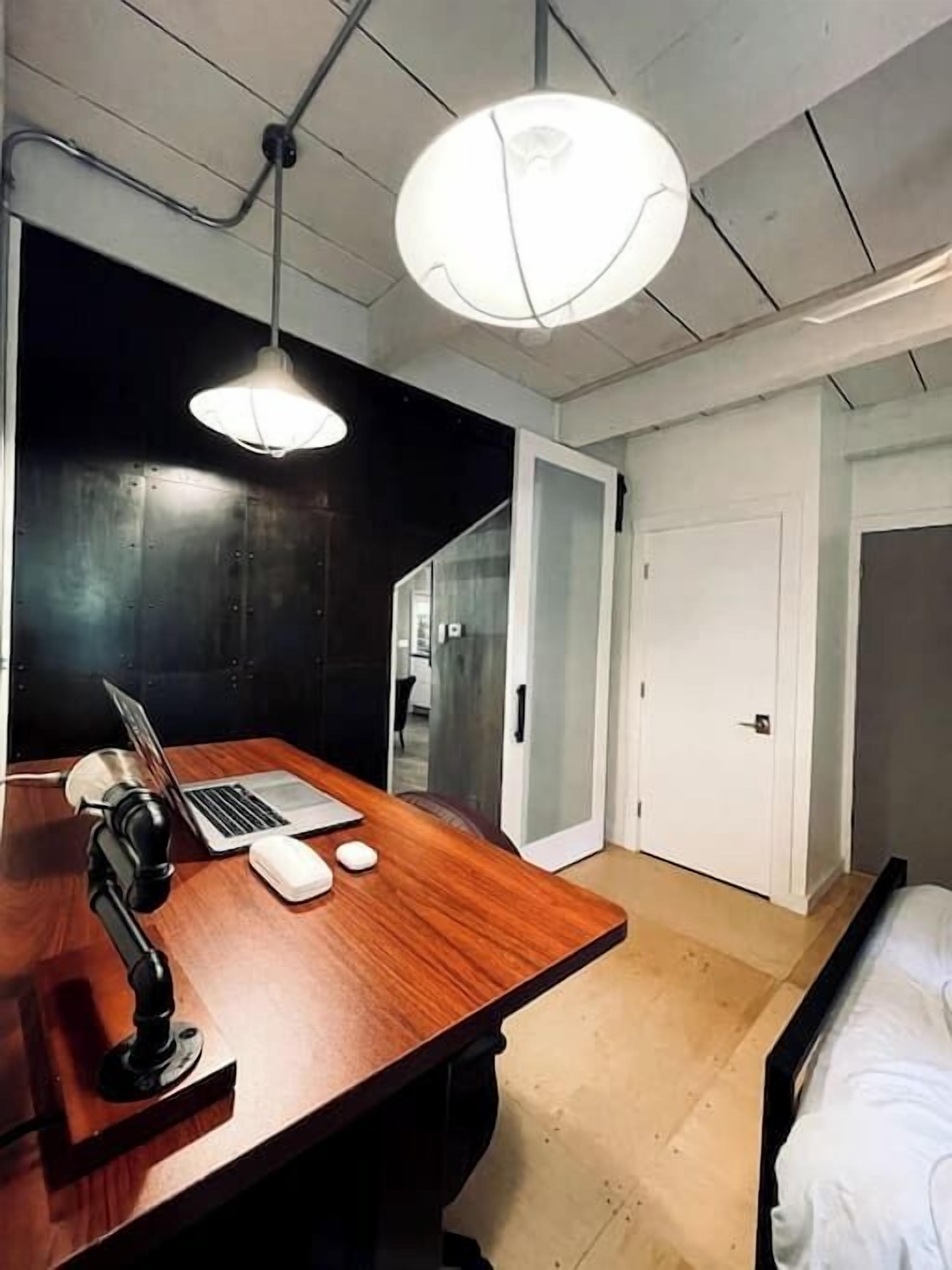 Una habitación con escritorio de madera, computadora portátil y lámpara de escritorio; una cama con ropa blanca; y una puerta que da a otra habitación.