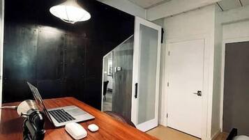 Una habitación con escritorio de madera, computadora portátil y lámpara de escritorio; una cama con ropa blanca; y una puerta que da a otra habitación.
