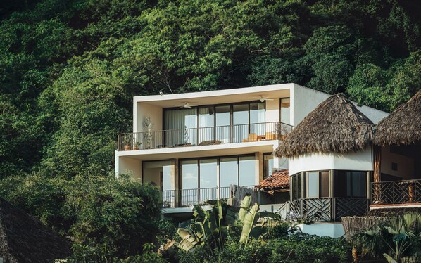 A modern two-story building with a balcony, surrounded by lush greenery.