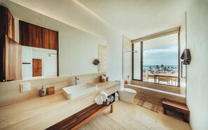 A modern bathroom with a large mirror, two sinks, a toilet, and a bench. A large window offers a view of the beach.