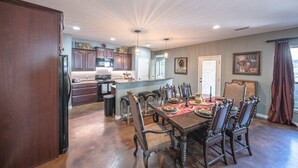 A spacious dining area with a wooden table set for six, surrounded by ornate chairs. A kitchen with modern appliances and wooden cabinets is visible in the background. - Right At Home-Midland Monterrey House (Midland)