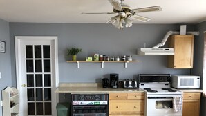 Cozinha com fogão branco, micro-ondas e cafeteira. Há um ventilador de teto, uma janela com moldura branca e uma porta com painel de vidro.