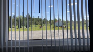 A window with vertical blinds, a view of a rural landscape with a field, trees, and a small building. - Restwell Motel Moncton (Berry Mills)