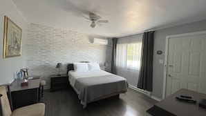 Quarto de hotel com cama, escrivaninha, cadeira, ventoinha no teto, janela com cortinas e porta.
