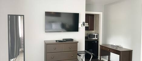 A modern living room with a flat-screen TV, a wooden dresser, a desk, and a kitchen area with a microwave.