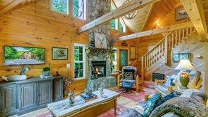 A cozy log cabin interior with a stone fireplace, wooden furniture, and a staircase leading to an upper level. - WindFall-Stunning, Pet Friendly, Creekside Cabin (Maggie Valley)