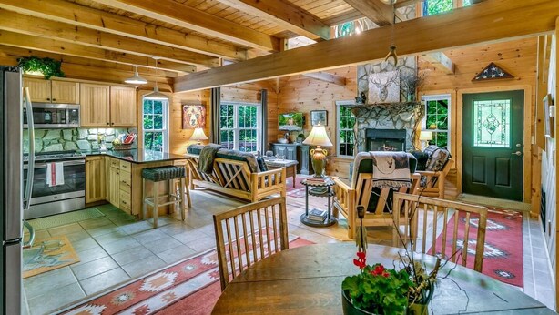 A spacious cabin with a kitchen, dining area, and living room. It features wooden beams, a stone fireplace, and large windows. - WindFall-Stunning, Pet Friendly, Creekside Cabin (Maggie Valley)