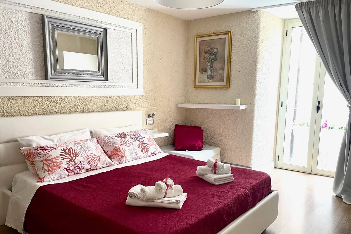 Een net opgemaakt bed met een rode sprei, witte kussens en een rood kussen op het nachtkastje. Daarboven hangt een spiegel in een lijst en aan de muur een schilderij.