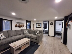Un salon moderne avec un canapé d’angle gris, une table basse en bois et un petit téléviseur. Une porte donne accès à une chambre à coucher équipée d’un lit et d’une armoire.